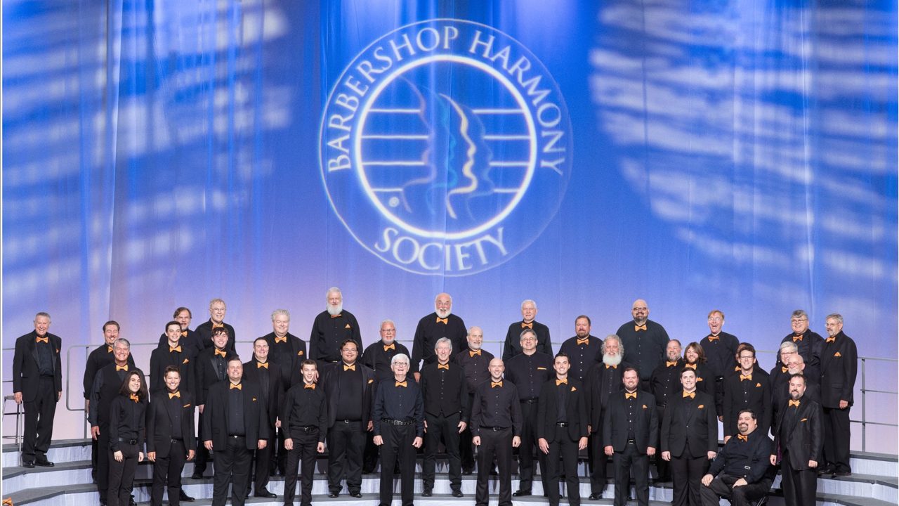 The chorus in black suits and orange bow ties standing on risers on the Barbershop Harmony Society International stage in Charlotte, 2022.