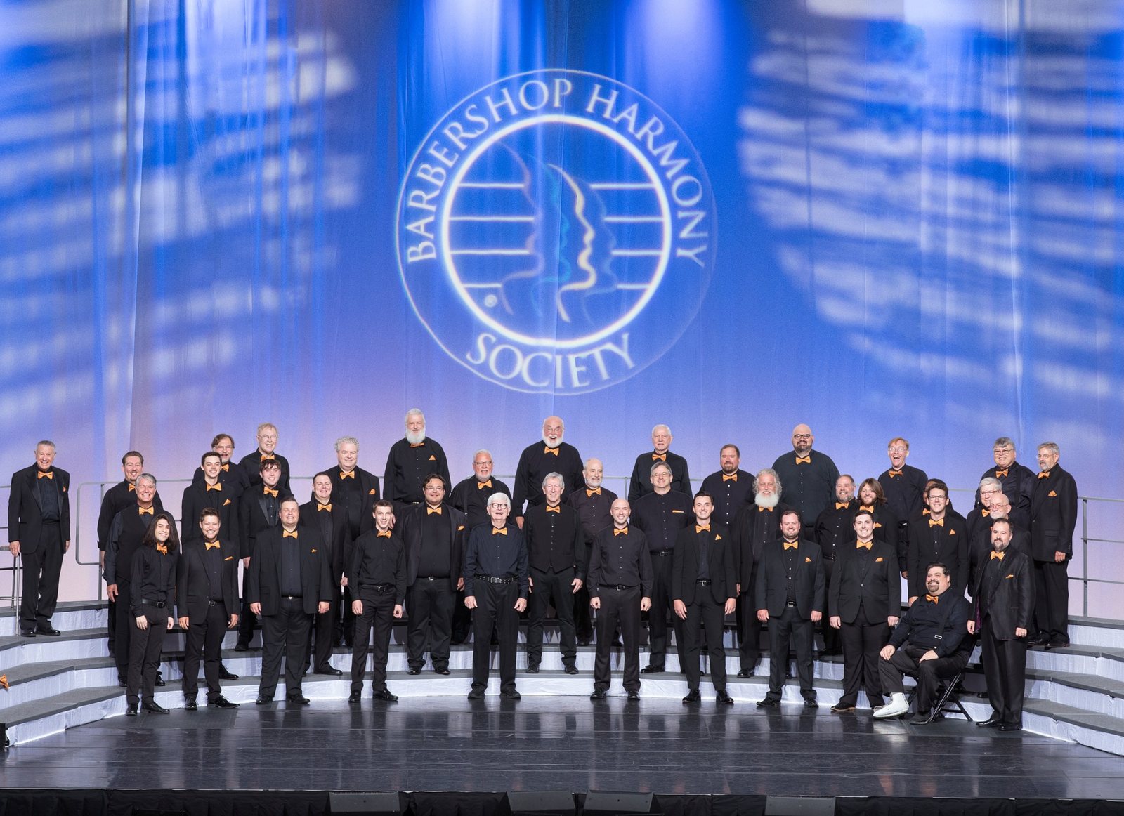 The chorus in black suits and orange bow ties standing on risers on the Barbershop Harmony Society International stage in Charlotte, 2022.