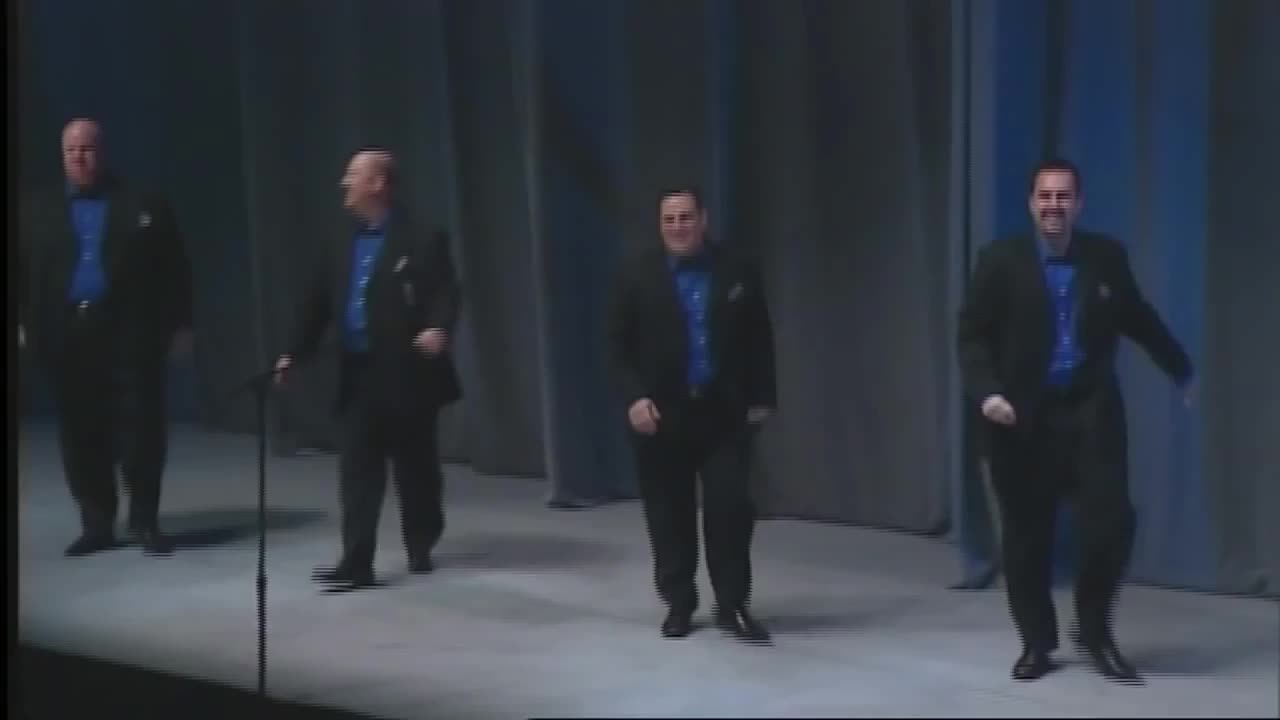 Video still: four men in black jackets and blue shirts stepping across a stage with blue curtains.
