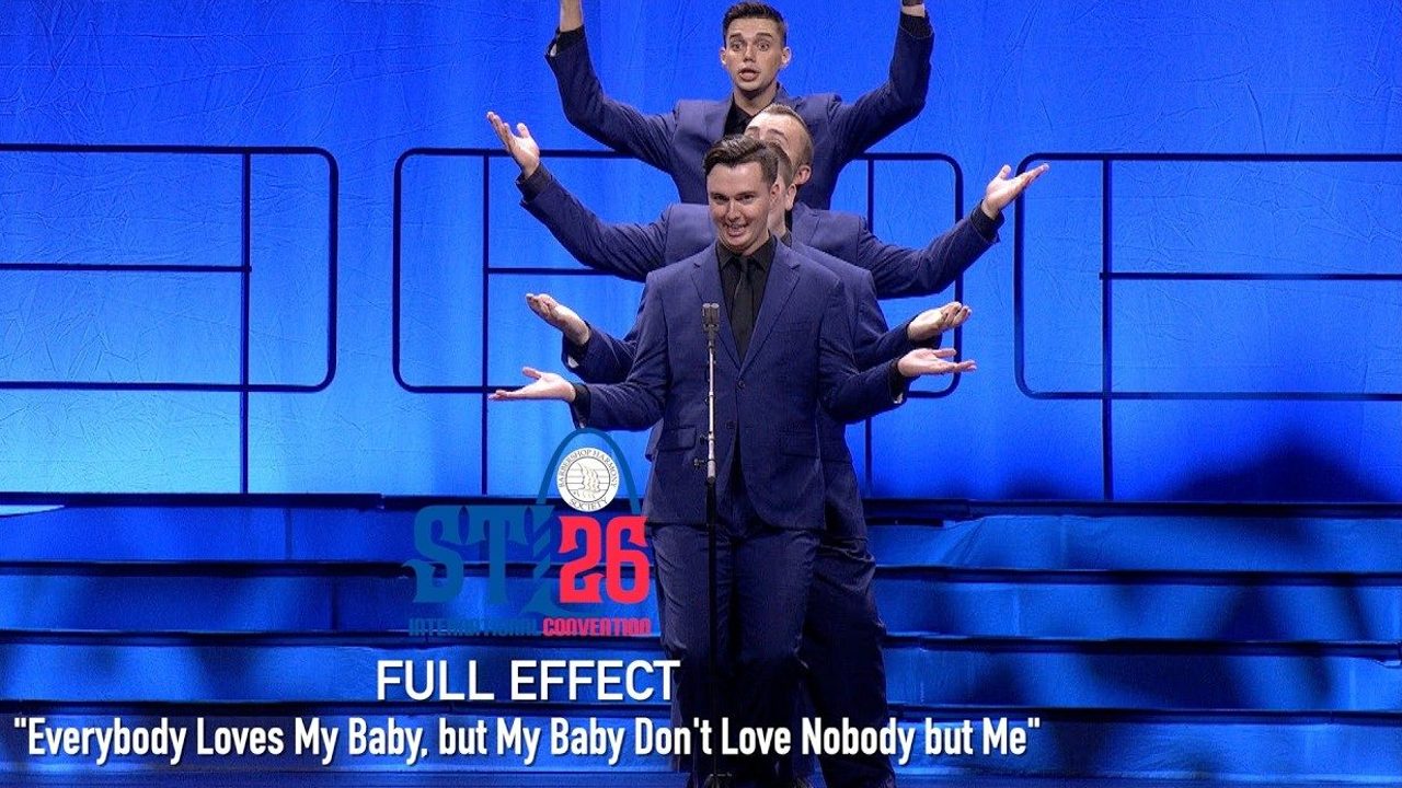 Video still: the four men of Full Effect in blue suits stacked one behind another at a microphone, arms out, on the St. Louis stage.