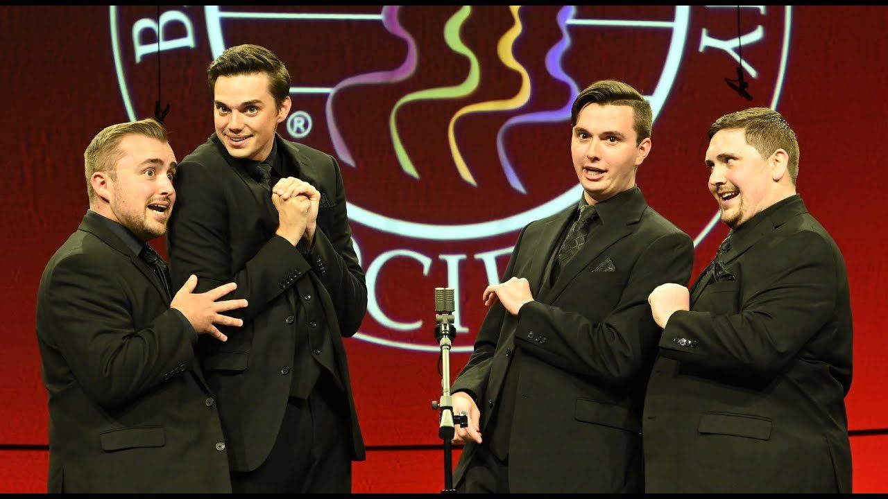 Video still: the four men of Full Effect in black suits leaning in around a microphone on the International stage in Denver, 2025.