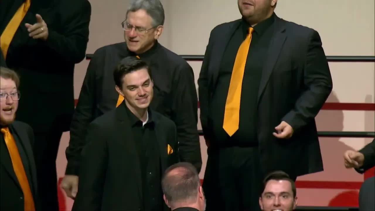 Video still from the chorus contest set: men in black with gold ties on the risers, one smiling at the camera.