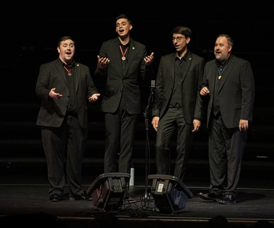 Step In Time, four men in dark jackets on a dark stage, three of them wearing medals.