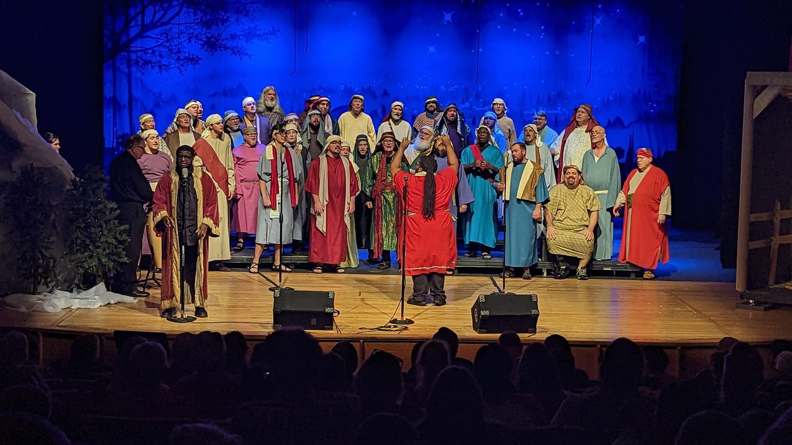 The chorus in robes on risers in front of a blue starry backdrop, with a singer in red facing them, at the 2022 Christmas show.