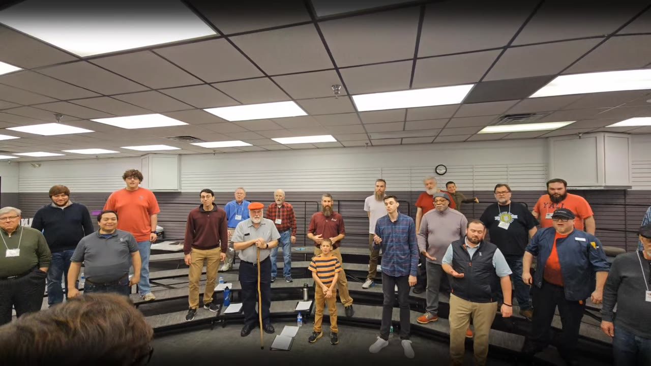 About forty men and a boy standing on risers in the rehearsal room, a man leading them, with his back to the camera.